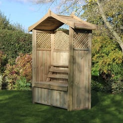 Natural wooden garden shed with a sloped roof, lattice panels, and bench seating, set in a lush backyard with trees and shrubs, ideal for outdoor storage or relaxing in your outdoor space.