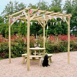 Natural wooden outdoor pergola with colorful bunting, picnic table, and a black puppy sitting on gravel in a lush garden setting. Perfect for backyard entertaining and garden decoration.