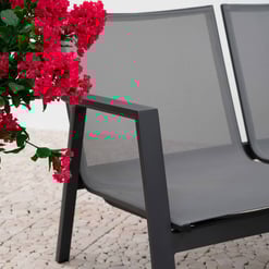 Modern outdoor patio bench with gray mesh seating and black metal frame, complemented by vibrant pink flowering plant for stylish garden and patio furniture display.