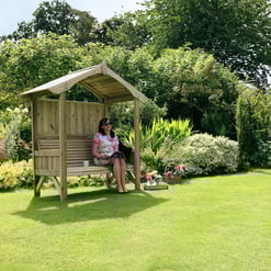 A woman relaxing on a wooden garden bench with a canopy in a lush, vibrant garden with colorful blooming flowers, greenery, and a sunny outdoor setting, ideal for outdoor living and garden decor.
