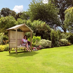 Vibrant garden with a wooden gazebo, lush greenery, colorful flowers, and two women relaxing outdoors in a spacious, well-maintained lawn, showcasing outdoor living and garden design.