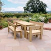 Natural wooden outdoor dining table with benches on a stone patio surrounded by vibrant garden flowers in a scenic countryside setting.