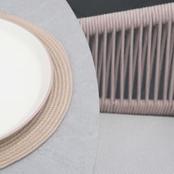 Minimalist round table with a modern, textured beige placemat, paired with woven wicker chairs, showcasing stylish outdoor furniture and home decor elements for a contemporary garden or patio setting.