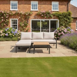 A stylish outdoor patio with modern seating set, lush flowering bushes, and a brick home background, perfect for outdoor living and garden relaxation.