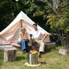 Cozy outdoor camping scene with a canvas tent, two friends sitting on cushions, and a small fire pit in a lush green setting, showcasing outdoor living gear from Empire Home and Garden.