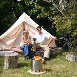 Cozy outdoor camping scene with a canvas tent, two friends sitting on cushions, and a small fire pit in a lush green setting, showcasing outdoor living gear from Empire Home and Garden.