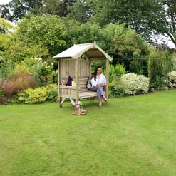 A woman relaxing on a wooden garden bench with a canopy, surrounded by lush, colorful plants and flowers in a well-maintained backyard garden.