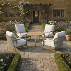 Elegant outdoor seating area with comfortable white cushioned chairs around a wooden table, set on a charming brick patio amidst lush garden flowers and a rustic stone house backdrop.