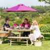 Family outdoor dining on a wooden picnic table with wine, under a large pink patio umbrella in a lush garden setting.