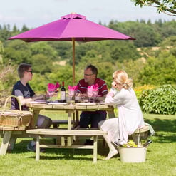 Family outdoor dining on a wooden picnic table with wine, under a large pink patio umbrella in a lush garden setting.