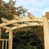 Natural wooden garden arbor with curved top arch, ideal for outdoor landscaping and welcoming yard entrances, surrounded by greenery and blooming plants.