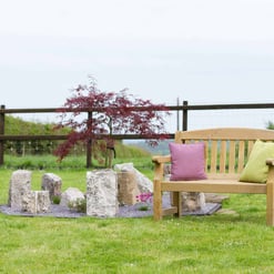 Lush green backyard with wooden garden bench adorned with pastel cushions, surrounding decorative rocks, a vibrant red Japanese maple tree, enclosed by a rustic wooden fence for outdoor relaxation.