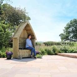 A person sitting on a wooden outdoor bench with a small shelter, surrounded by lush greenery and colorful plants, under a bright blue sky - perfect for garden relaxation and outdoor furniture.