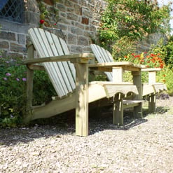 Rustic wooden outdoor garden bench with armrests, situated beside colorful flowering plants and lush greenery on a gravel pathway. Perfect for garden seating and relaxation.