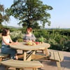 Natural outdoor wooden picnic table set in a scenic garden with two women enjoying drinks amidst lush greenery and blooming flowers, perfect for outdoor dining and garden furniture ideas.