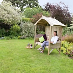 A woman relaxing on a wooden garden swing with a canopy, surrounded by lush greenery and colorful plants in a well-maintained garden.