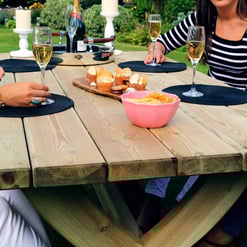 Enjoying a summer outdoor gathering with friends around a rustic wooden patio table, featuring snacks, drinks, candles, and tableware, in a lush garden setting.