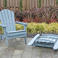 Colorful Adirondack chairs and footstools set on a stone patio in a landscaped garden with ornamental grasses and a wooden fence.