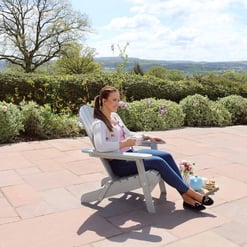 Relaxing woman sitting on a garden chair enjoying outdoor scenery on a sunny day with lush greenery and flowering bushes.