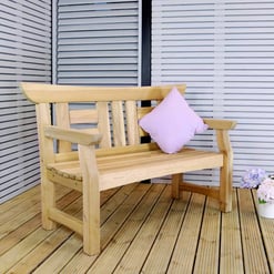 Natural wood outdoor garden bench with purple cushion on a wooden deck with potted flowers and slatted wall.