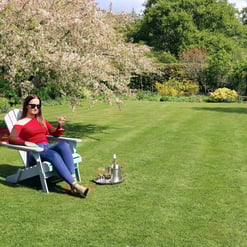 Relaxing woman outdoors on garden lawn, enjoying summer with wine and cocktails in a spacious backyard, surrounded by blooming trees and lush greenery, perfect for outdoor leisure and entertaining.
