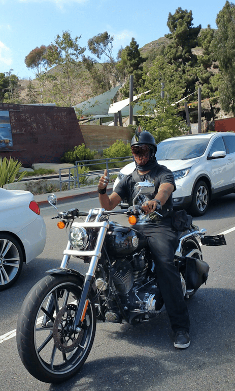 Karif Battle, founder of Theoretical Fiends LLC, is sitting on his Harley Davidson motorcycle in traffic, giving his camera man a thumbs up, dressed in dark supernatural afro gothic metal biker ring on each finger attire.