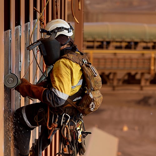 Side view of rope access technician welder services wearing white helmet head fall protection PPE, face shield abseiling working at height using power grinder grinding wall preparation prior weld