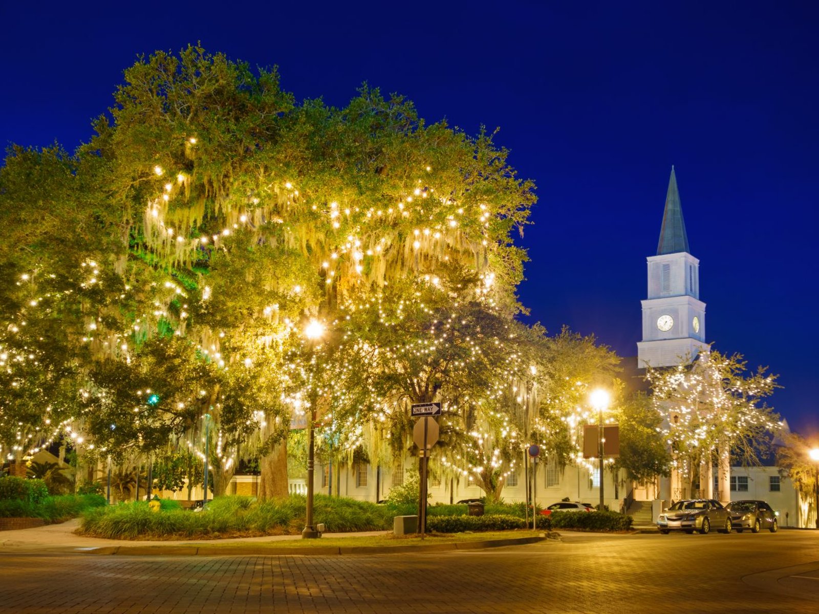 Large oak trees covered in glowing string lights beside a white church in downtown Tallahassee at night.