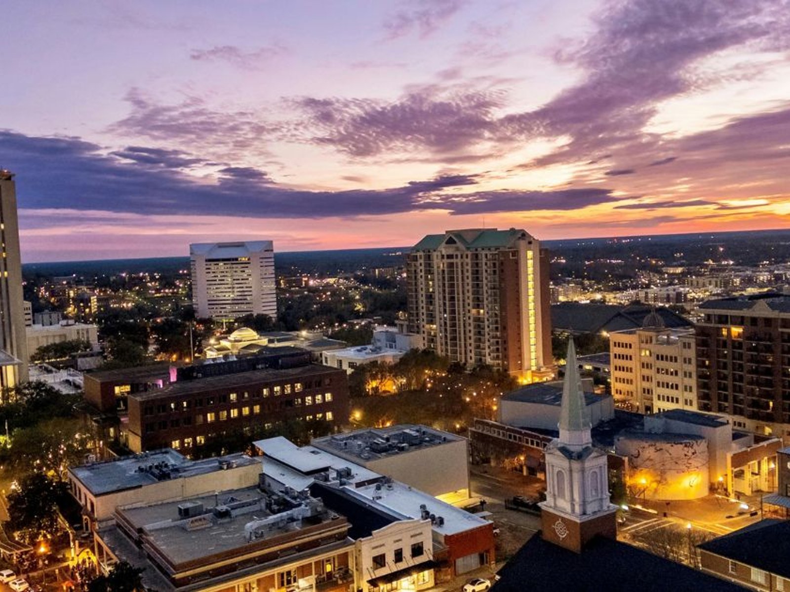 Aerial view of downtown Tallahassee’s buildings and illuminated streets beneath a colorful sunset sky.