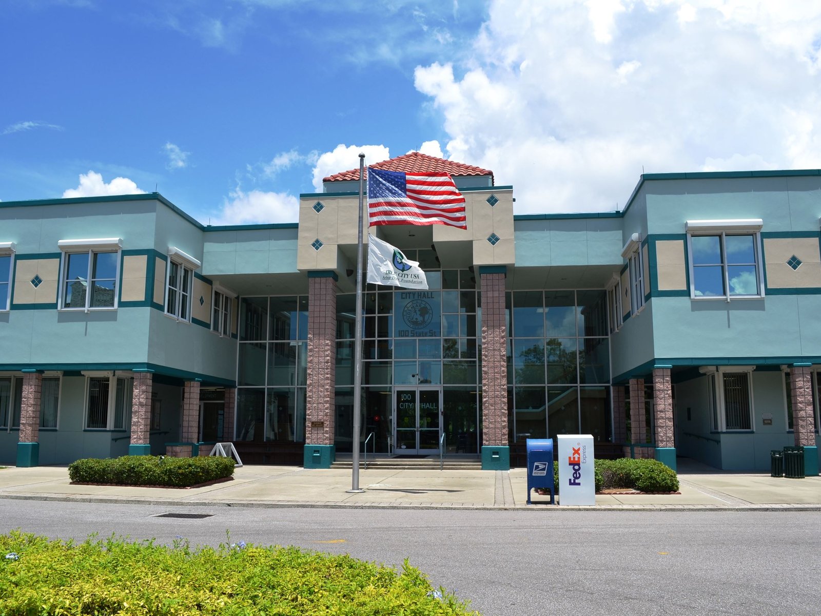 Front exterior of Oldsmar City Hall in Oldsmar, Florida, featuring a blue and beige building, glass entrance, palm trees, and American and city flags flying outside.