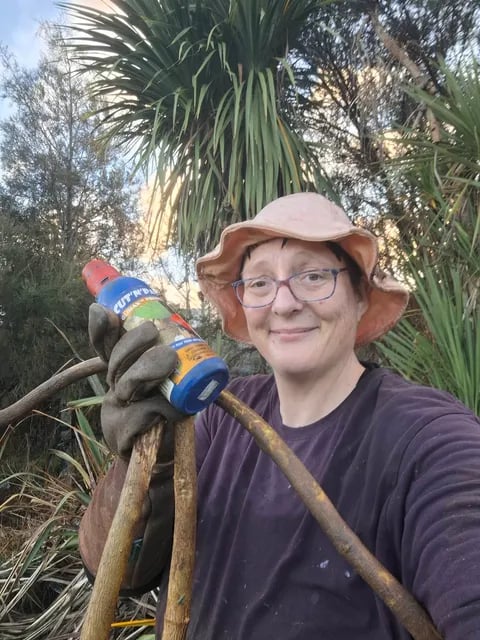 A smiling woman wearing glasses, a sun hat, and work gloves holding a bottle of weed gel and cut vine stems outdoors.