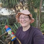 A smiling woman in a pink sun hat holding a bottle of weed gel and thick cut stems in front of native foliage.