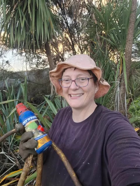 A smiling woman in a pink sun hat holding a bottle of weed gel and thick cut stems in front of native foliage.