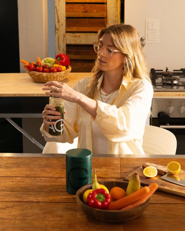 A woman mixing AG1 at a kitchen table beside a bowl of fruit
