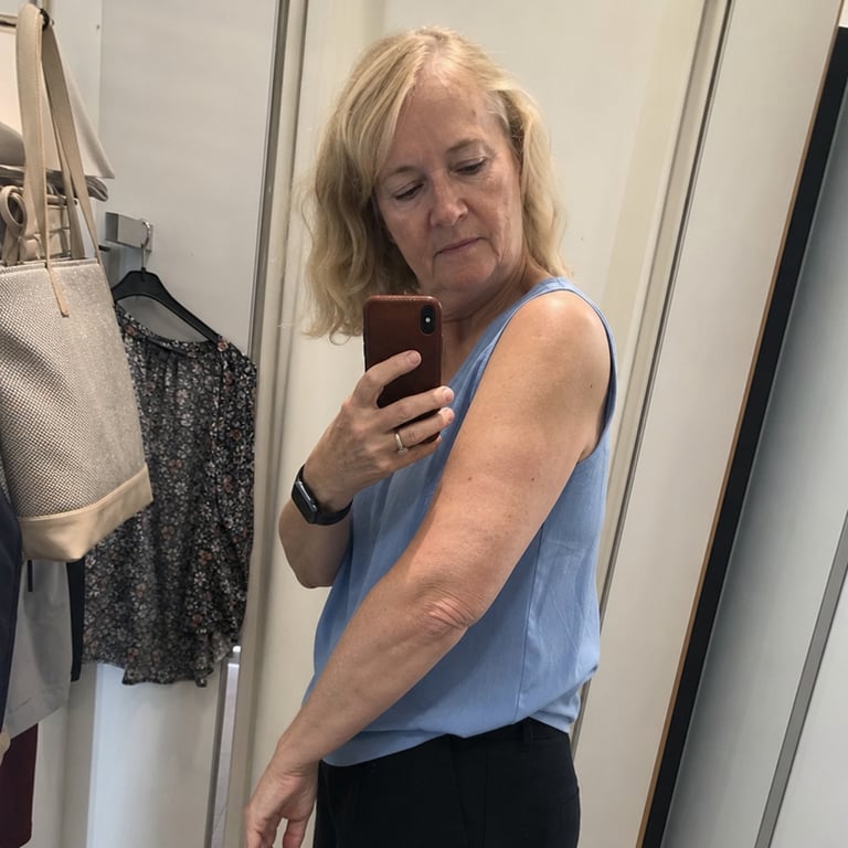 A woman in a changing room mirror wearing a sleeveless top, glancing at her upper arm