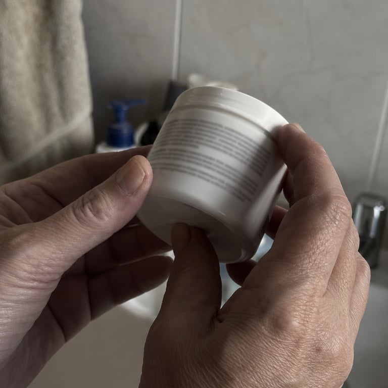 Mature hands holding a white cream tub turned over to read the ingredients label