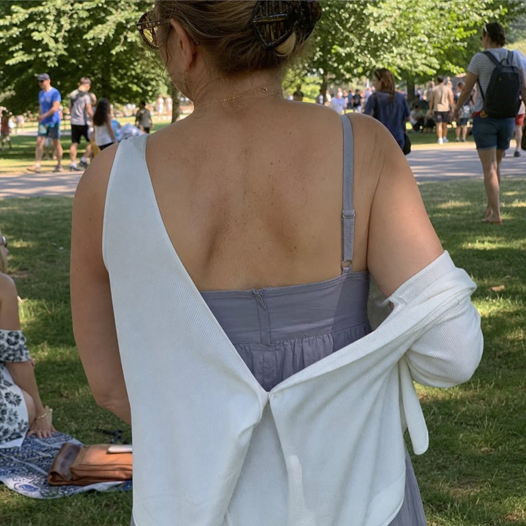 A woman pulling her cardigan sleeve down over her upper arm in a sunny park while others around her wear summer clothes