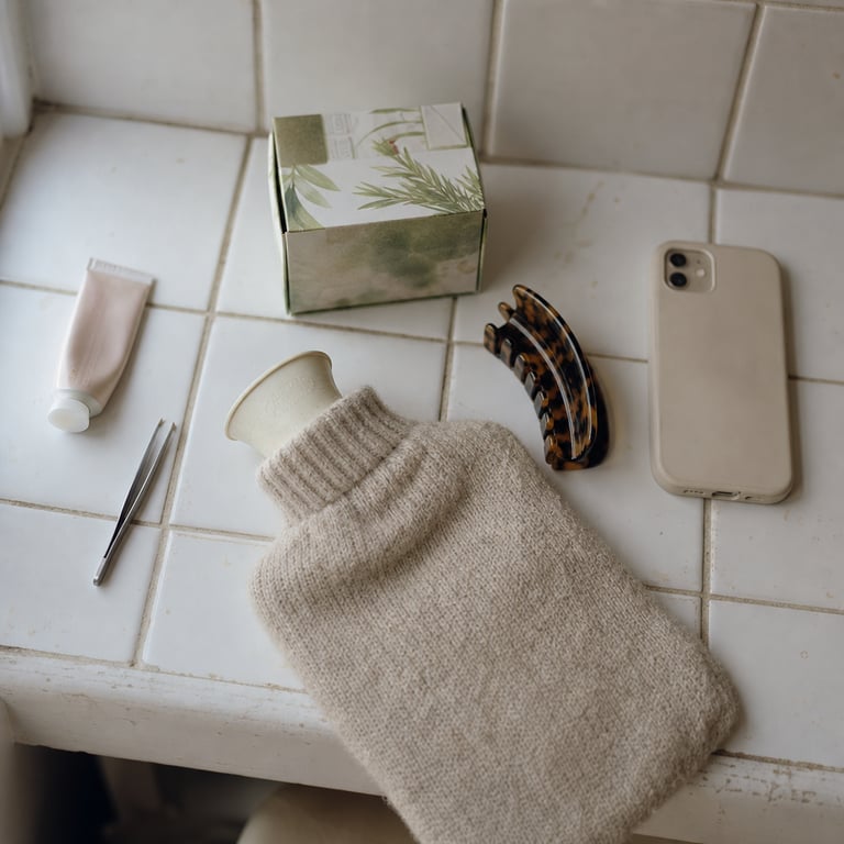 An overhead view of a bathroom shelf: spot treatment, tweezers, tea, a hot water bottle, a hair claw, a phone face down