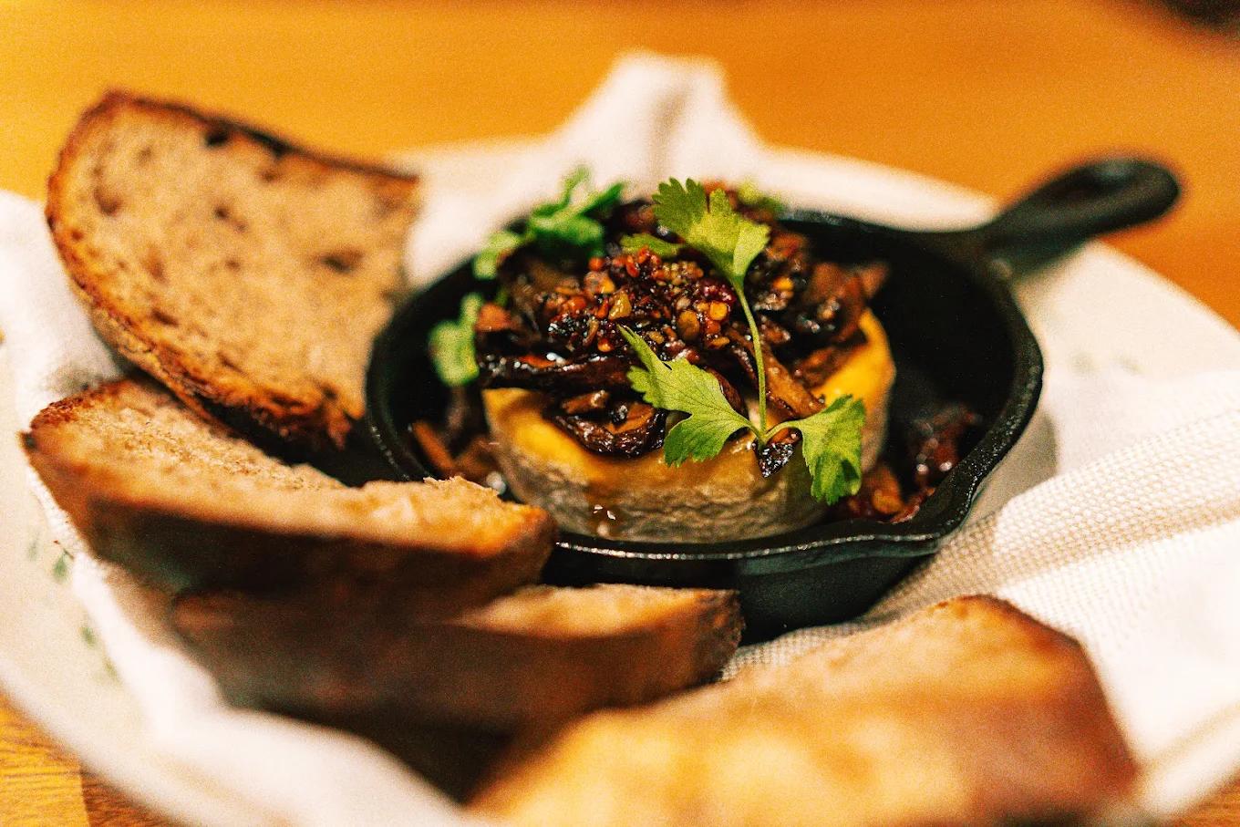 Slices of toasted bread surround a small cast-iron skillet filled with baked cheese and sautéed mushrooms.