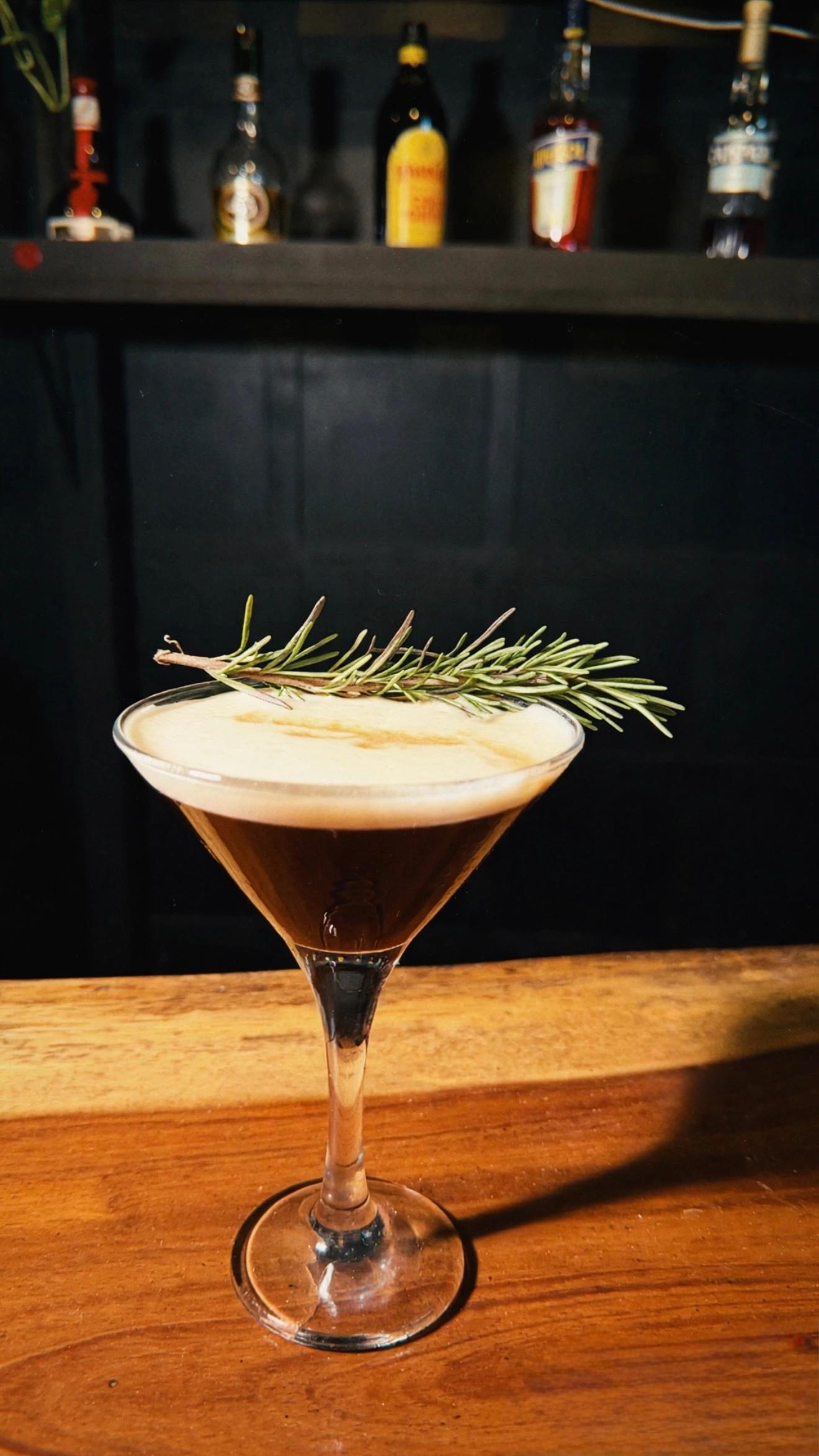 Cocktail in a martini glass garnished with rosemary, set on a wooden bar in a dim venue.