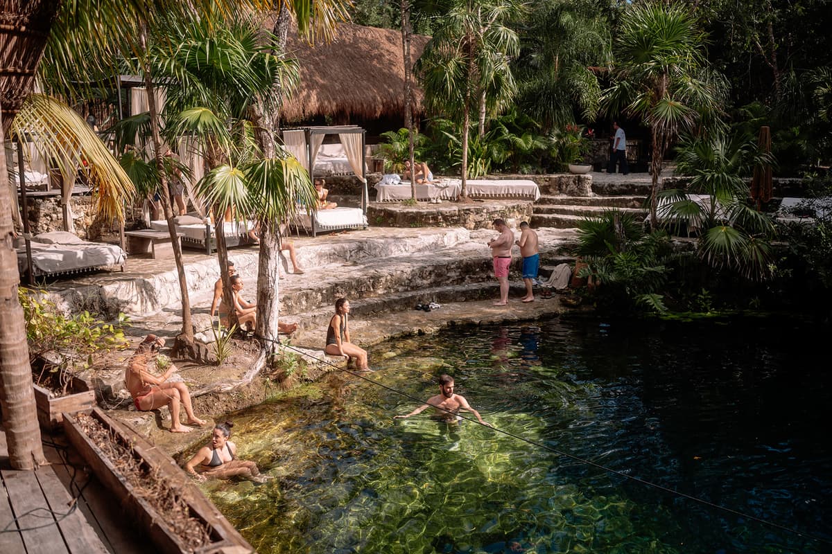 Guests swimming and relaxing in a clear cenote surrounded by stone platforms, palm trees, and thatched-roof structures at a