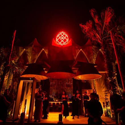 Bonbonniere Tulum glowing red at night, with palm trees, umbrellas, a bar and people gathered outside.