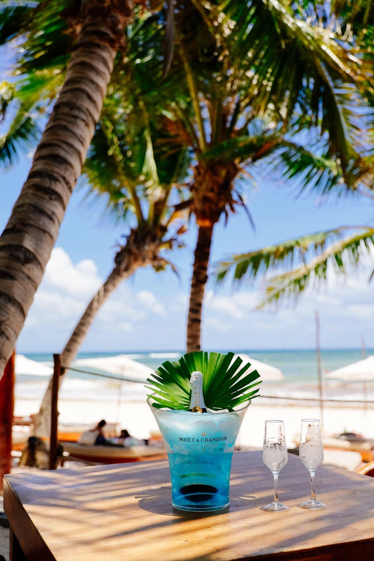 Tulumania beach club table with a blue champagne ice bucket and flutes overlooking palm trees and the sea.