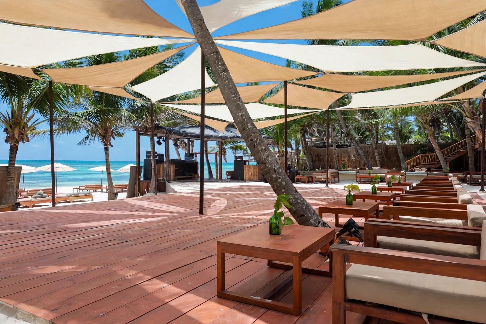Tulumania’s beachfront wooden deck has lounge seating, shade sails, palm trees, and a bright turquoise sea.