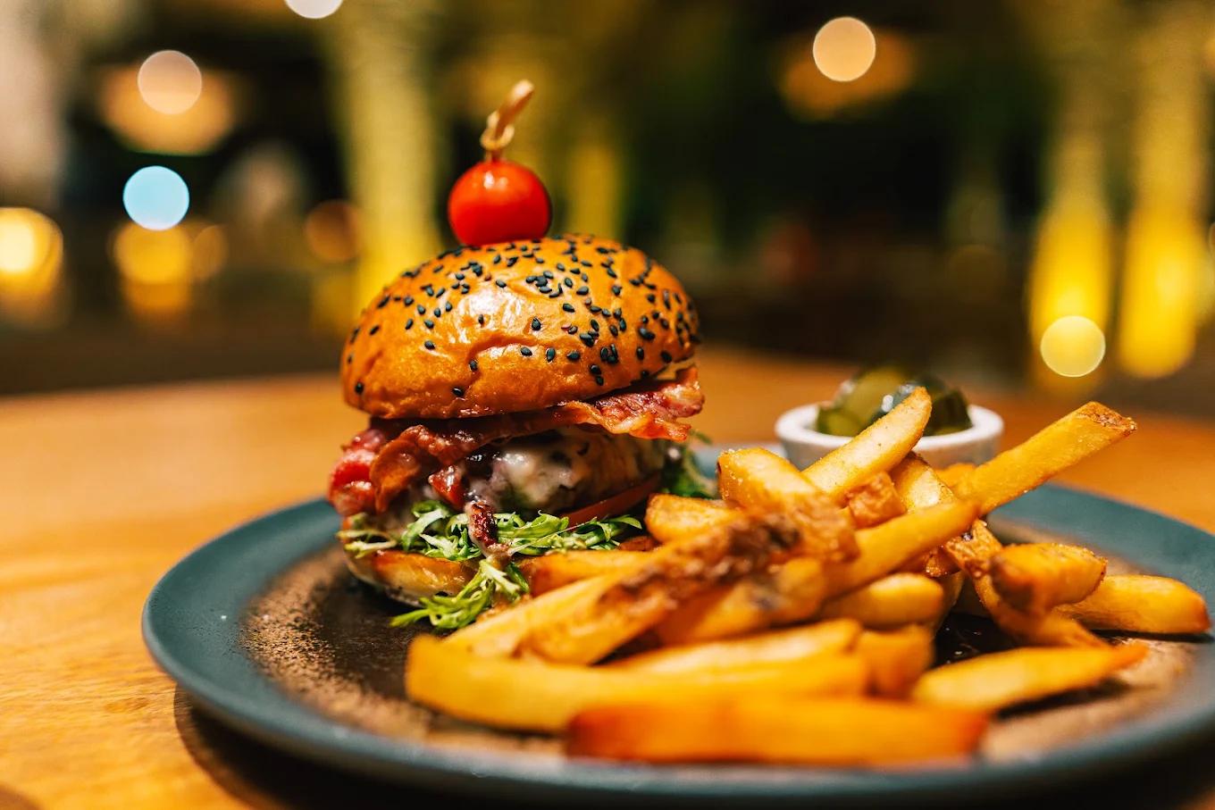 A bacon cheeseburger and french fries are served on a plate with warm, ambient lighting in the background at Atta.