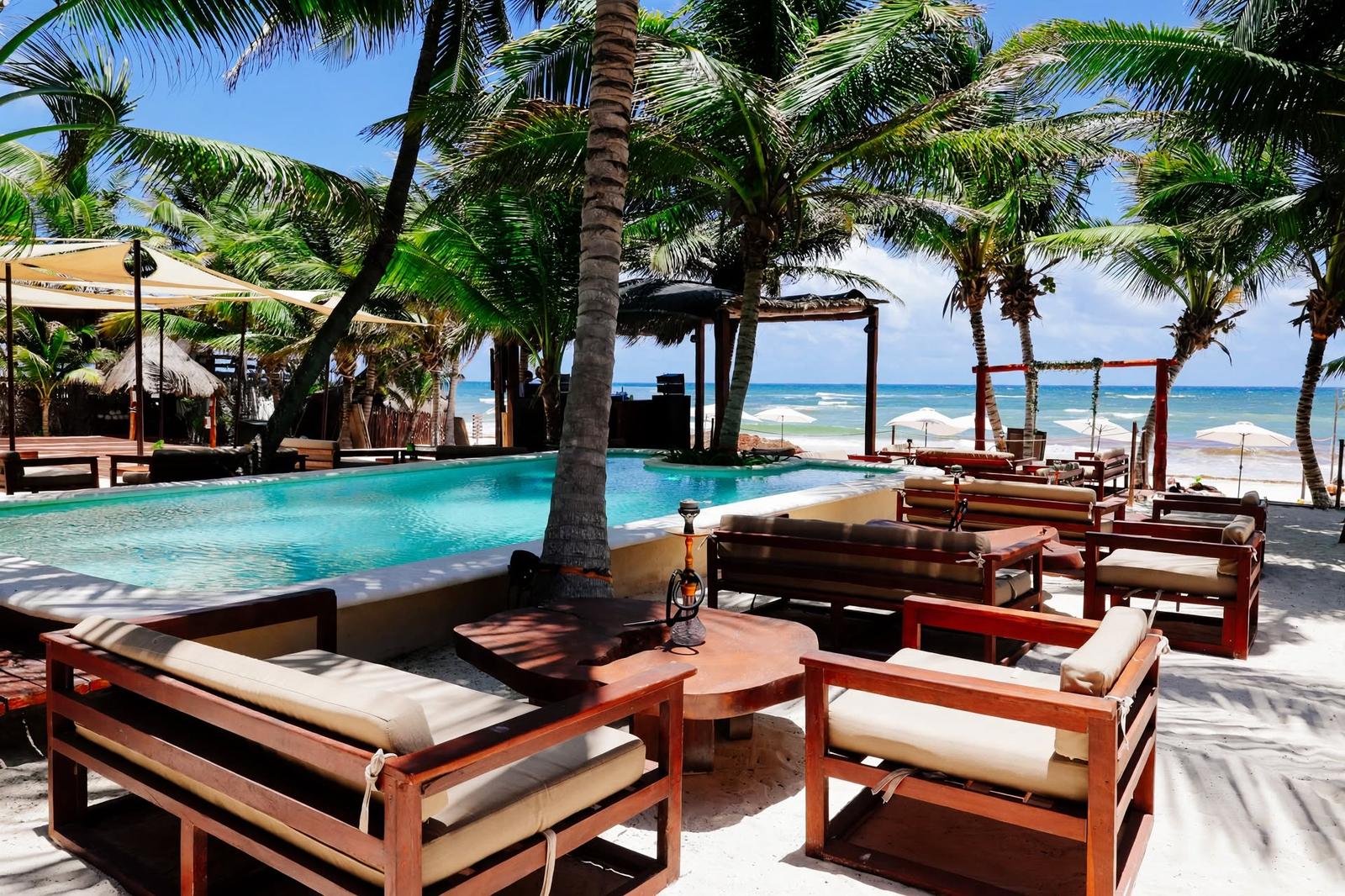 Tulumania’s poolside lounge overlooks a sandy beach and turquoise sea, shaded by palm trees.
