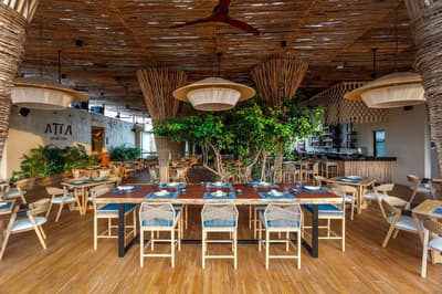 Atta's open-air dining room features wooden tables, woven chairs, lush greenery, and a thatched ceiling.