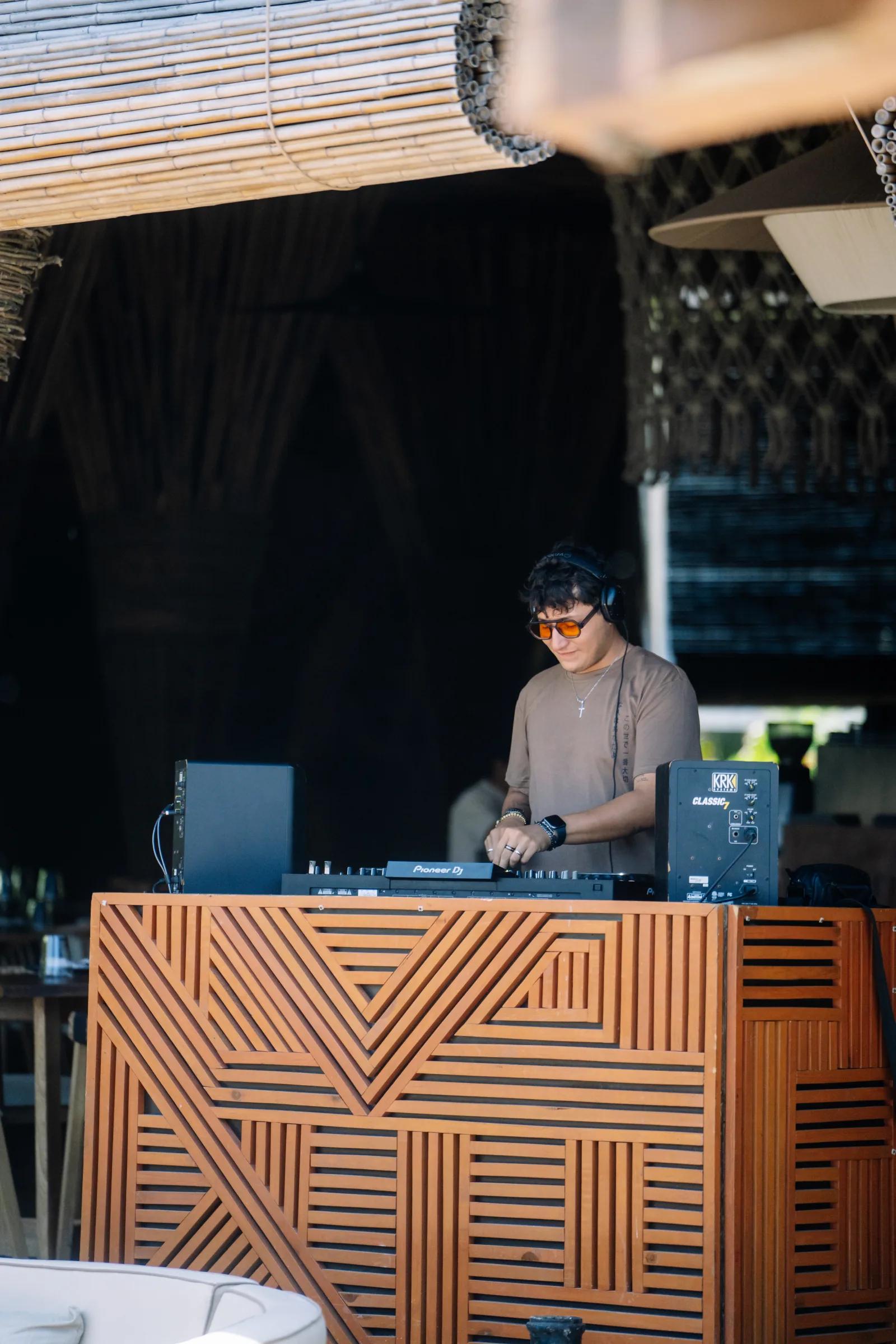 A DJ wearing orange sunglasses performs at the carved wooden DJ booth at Atta under a bamboo roof