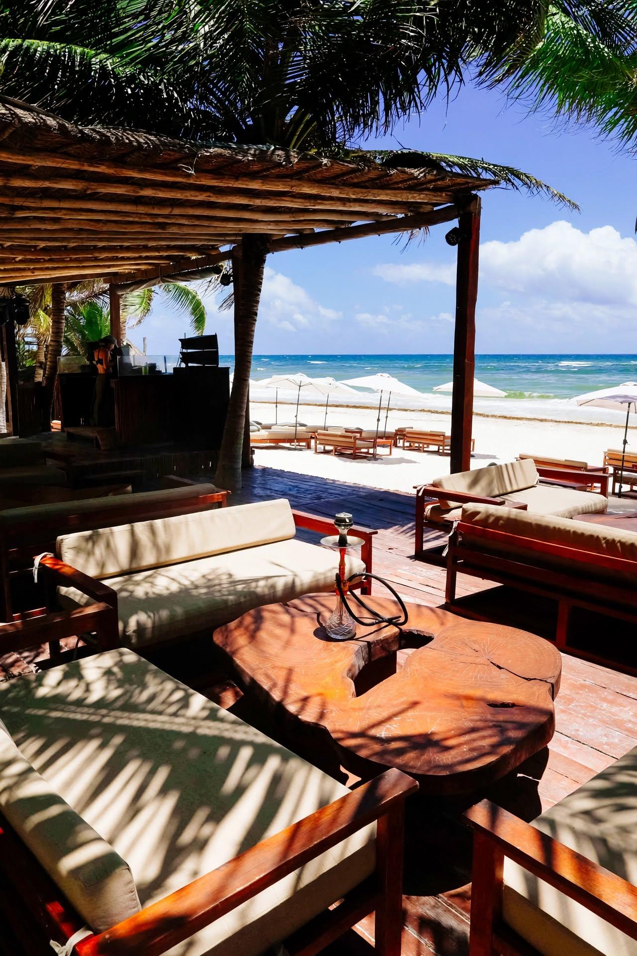 Tulumania’s beachfront deck has cushioned seating, a hookah, palm shade, umbrellas and ocean views in bright daylight.