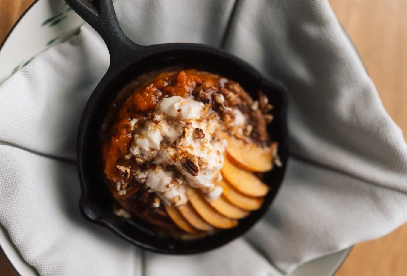 Sliced peaches, whipped cream, and chopped nuts top a warm dessert served in a small cast-iron skillet at